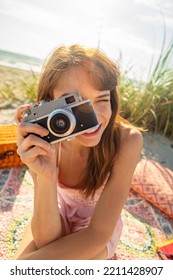 Smiling Caucasian Girl Enjoying Carefree Beach Vacation Sitting On Picnic Blanket With Camera Photographing Fun In The Sun