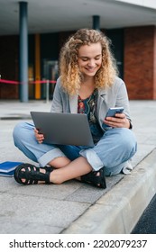 Smiling Caucasian Girl Sitting In A Street Working At The Computer While Reading Messages On Smartphone. Summer, Internet, Education, Campus And Teenage Concept