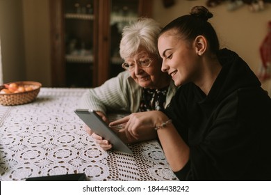 A smiling caucasian grandmother and her teenage granddaughter look at a tablet while sitting at a table in the kitchen. The granddaughter teaches her grandmother to use new technology - Powered by Shutterstock