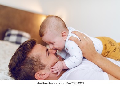 Smiling Caucasian Handsome Dad Lying On Bed In Bedroom And Holding His Loving Adorable Six Months Old Son On His Chest. Baby Biting Dad's Nose. Unconditional Love Concept.