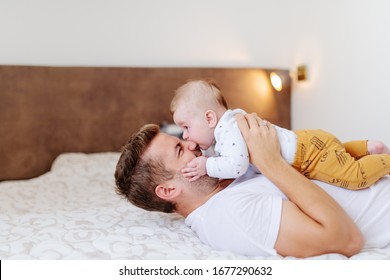 Smiling Caucasian Handsome Dad Lying On Bed In Bedroom And Holding His Loving Adorable Six Months Old Son On His Chest. Baby Biting Dad's Nose. Unconditional Love Concept.