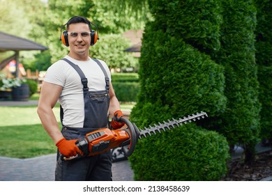 Smiling caucasian man in overalls, safety glasses and gloves holding electric hedge trimmer while posing at summer garden. Modern gardening tool for seasonal work.  - Shutterstock ID 2138458639