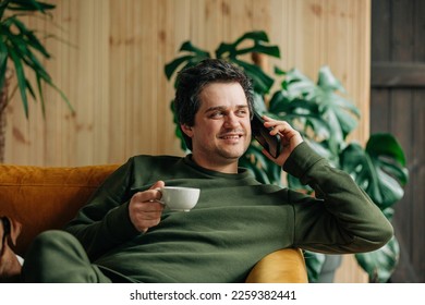 Smiling caucasian man with smartphone and cup of coffee sits in sofa at home - Powered by Shutterstock