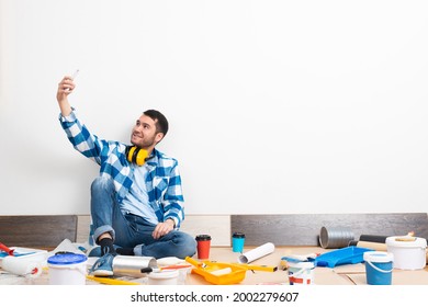 Smiling Caucasian Man Taking Selfies Photo On Mobile Phone While Sitting Near Wall. House Remodeling And Interior Renovation. Young Bearded Guy Sitting On Floor Among Construction Tools At Home.