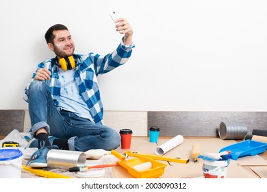 Smiling Caucasian Man Taking Selfies Photo On Mobile Phone While Sitting Near Wall. House Remodeling And Interior Renovation. Young Bearded Guy Sitting On Floor Among Construction Tools At Home.