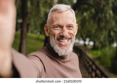 Smiling caucasian mature man looking at the camera, taking selfie on smart phone, having video call, vlogging blogging outdoors in park. - Powered by Shutterstock