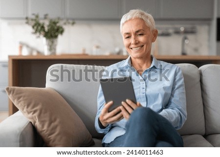 Smiling caucasian mature woman using digital tablet, sitting with computer gadgets while relaxing on sofa at modern home interior on weekend. Lady browsing websites and internet indoor