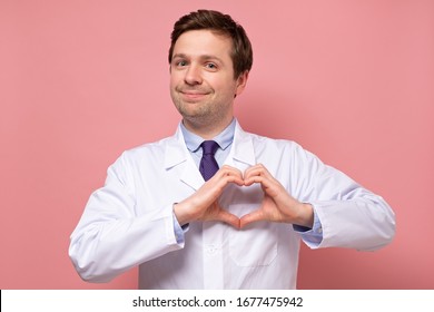 Smiling caucasian medical male doctor showing heart figure with fingers over pink background