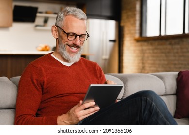 Smiling caucasian middle-aged mature man in glasses using digital tablet for e-learning, paying bills online, e-commerce, remote work, social media online at home - Powered by Shutterstock