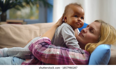 Smiling Caucasian Mother Holding Adopted Mixed-race Baby At Home. Side View Of Foster Mother Lying On Couch With Adorable Infant At Home. Babysitter Playing With Little Child