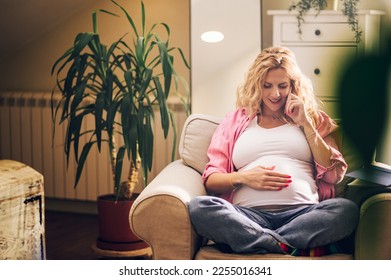 Smiling caucasian pregnant woman using smartphone while resting on cozy chair in a living room at home and making preparations for an upcoming child online. Talking on a mobile phone. Copy space. - Shutterstock ID 2255016341