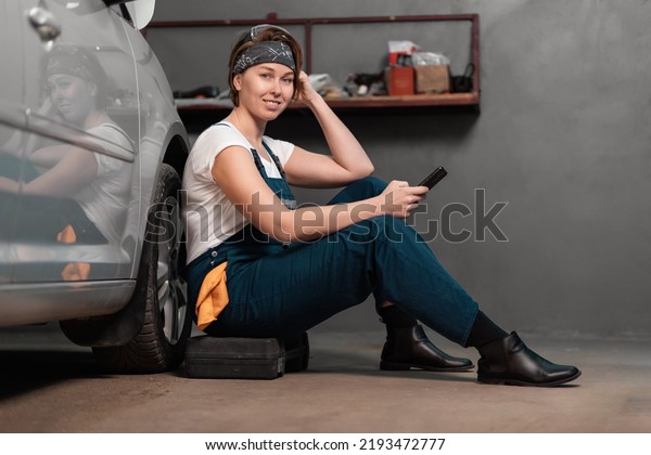 Smiling caucasian pretty woman\
mechanic is sitting leaning on a car in an auto repair shop and\
using cellphone. The concept of women\'s work in male\
professions.