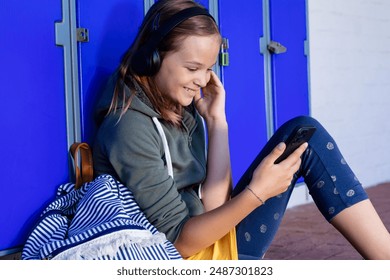 Smiling caucasian schoolgirl in headphones using smartphone sitting by lockers at school, copy space. Education, inclusivity, childhood, technology, elementary school and learning concept. - Powered by Shutterstock