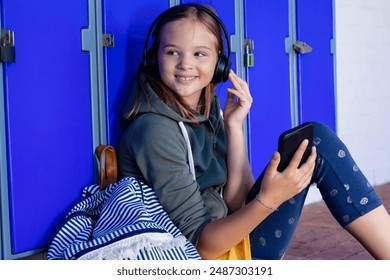 Smiling caucasian schoolgirl in headphones using smartphone sitting by lockers at school, copy space. Education, inclusivity, childhood, technology, elementary school and learning concept. - Powered by Shutterstock