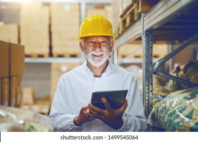 Smiling Caucasian senior adult employee in white uniform and with yellow helmet on head checking on goods in warehouse by using tablet. - Powered by Shutterstock