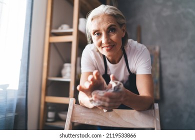 Smiling caucasian senior female master wiping hands with rag after molding clay in art studio. Small business and entrepreneurship. Home hobby and leisure. Woman wear work apron and look at camera - Powered by Shutterstock