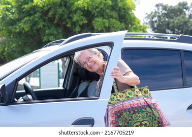 Smiling Caucasian Senior Woman Entering In Her Car, Happy Grandmother With Large Handmade Bag Open The Door To Enter In The Vehicle Ready To Drive