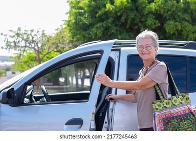 Smiling Caucasian Senior Woman Entering In Her Car, Happy Grandmother With Large Handmade Bag Open The Door To Enter In The Vehicle Ready To Drive