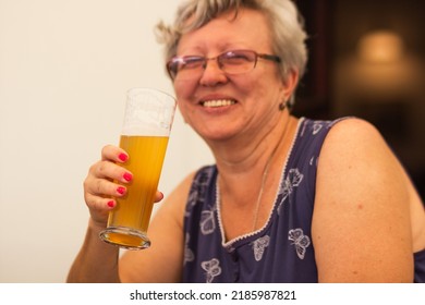 Smiling caucasian senior woman with gray hair holding and drinking glass of light beer in craft czech brewery. Alcoholic drink. Weekend meeting with friends in pub. Oktoberfest concept.