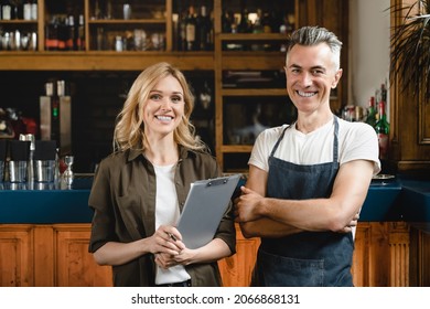 Smiling caucasian small business owner waiter barista with female auditor holding clipboard standing at the bar restaurant cafe counter checking products quality looking at the camera - Powered by Shutterstock