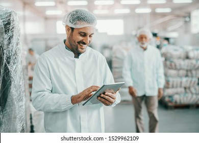 Smiling Caucasian Supervisor In Sterile Uniform Standing In Food Factory With Tablet In Hands And Checking Quality Of Goods.