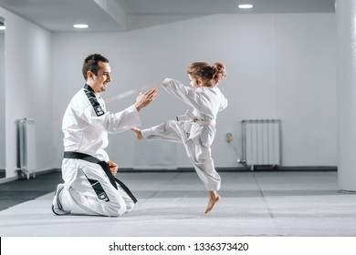 Smiling Caucasian taekwondo trainer in dobok kneeling and holding hand up while little girl kicking him.