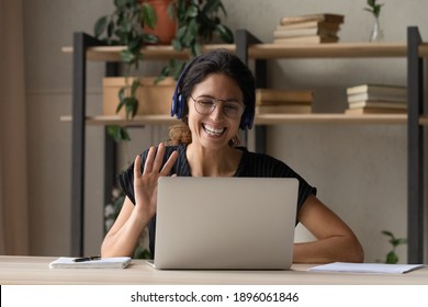 Smiling Caucasian Woman In Headphones Wave Greet To Camera Talking On Video Call On Laptop. Happy Young Female In Glasses And Earphones Have Webcam Digital Conference Or Meeting On Computer.