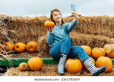 Smiling caucasian woman sitting on straw bales and holding pumpkin and making selfie on phone. Selecting Thanksgiving and Halloween holidays decor on agriculture farm. Autumn fall festive mood