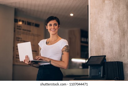Smiling caucasian woman standing in her coffee shop with a laptop. Female manager with laptop in a restaurant.