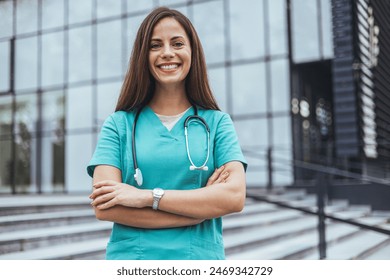 A smiling Caucasian woman in teal scrubs stands confidently with arms crossed outside a medical building, stethoscope around her neck and watch on her wrist, symbolizing professional healthcare. - Powered by Shutterstock