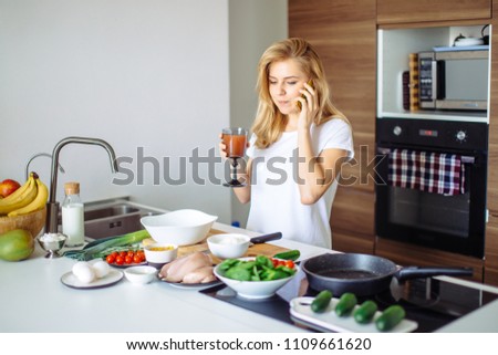 Smiling caucasian woman while making healthy meal, drinking smoothie and talking with a friend at phone in the domestic kitchen.