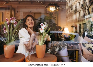 Smiling Caucasian Young Brunette Woman Chooses Bouquet In Flower Shop. Model Wearing White Sundress Spends Her Leisure Time Summer. Enjoying Little Things, Positive Attitude Concept