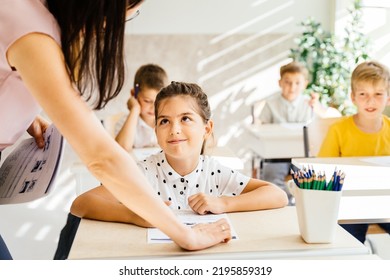 Smiling Caucasian Young Female Teacher Teaching Nice American Elementary Girl At Desk In Classroom. Education And School Concept.