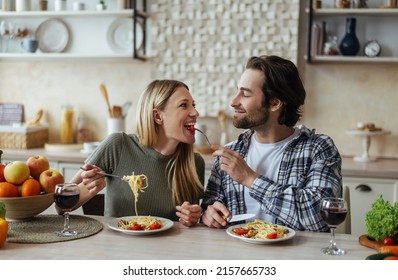 Smiling Caucasian Young Male With Stubble Feeds Blonde Woman, Couple Have Fun Together In Light Kitchen Interior. Romantic Dinner With Wine At Home. Love, Relationship, People And Family, Covid-19