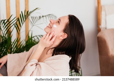 Smiling Caucasian Young Woman Applying Cream On Face At Home. Morning Beauty Routine. Skincare Concept