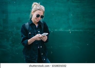 Smiling caucasian young woman sending message on smartphone standing near promotional copy space background, positive trendy hipster girl blogging in social network using mobile phone with internet - Shutterstock ID 1018031032