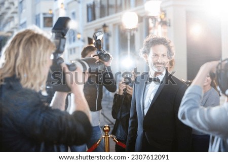 Smiling celebrity in tuxedo being photographed by paparazzi at red carpet event