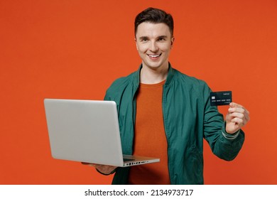 Smiling Charismatic Happy Young Brunet Man 20s Wears Red T-shirt Green Jacket Using Laptop Pc Computer Hold Credit Bank Card Doing Online Shopping Isolated On Plain Orange Background Studio Portrait