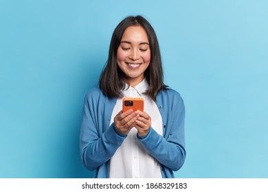 Smiling charming brunette Asian woman uses mobile phone happy texting in social networks addicted to modern technologies wears casual jumper isolated over blue background. Online communication - Shutterstock ID 1868329183