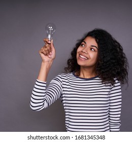 Smiling charming cute young dark-skinned girl with curly gorgeous hair, in a striped casual sweater, holds a light bulb in her right hand, smiles and looks at her with an idea.