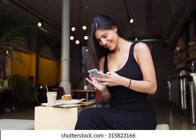 Smiling charming latin woman read good news on her mobile phone during breakfast in cafe terrace in summer day, young female student typing message on cell telephone while sitting in restaurant