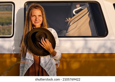 Smiling Charming Woman Posing Next To Retro Old Van, Holding Black Hat In Hands, Looking Posing At Camera. Outdoor Portrait. At Sunset, Summer Holidays In Nature On Road. Alternative Travel Concept