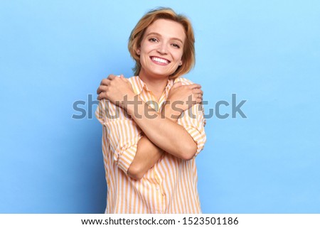 smiling charming woman putting her palms on shoulders posing to the camera. romantic girl embracing herself, close up portrait, isolated blue background