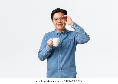 Smiling Cheerful Asian Guy, Office Worker In Glasses Having Lunch Break, Standing With Cup Of Coffee Over White Background. Man Drinking From Mug And Looking Upbeat, Morning Routine