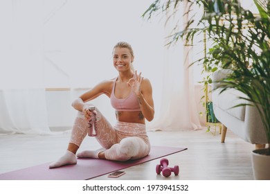 Smiling cheerful attractive young strong sporty fitness woman wearing pink tracksuit doing yoga exercises sitting holding bottle of water showing OK gesture stretching on mat floor at home gym indoor