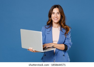 Smiling cheerful beautiful attractive young brunette woman 20s wearing basic jacket standing working on laptop pc computer looking camera isolated on bright blue colour background studio portrait