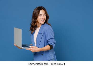 Lächelnd fröhlich schöne junge brunette Frau 20er Jahre, die eine einfache Jacke trägt, die auf Laptop PC-Computer arbeitet und abseits auf hellblauem Hintergrund Studioaufnahme steht