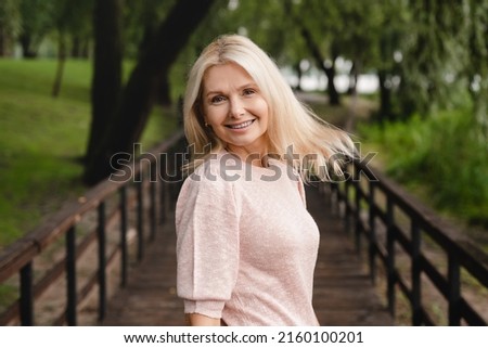 Smiling cheerful beautiful blonde caucasian mature woman looking at the camera while walking in city public forest wood park Foto stock © 