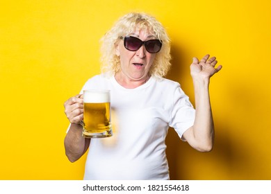 Smiling Cheerful Blonde Old Woman In A White T-shirt Holds A Glass Of Beer And Waves Her Hand On A Yellow Background