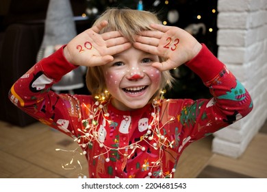 smiling cheerful child in festive make  up deer  numbers new year 2023 are written palms  The kid is waiting for miracle Christmas Eve  New Year's winter holidays  pampering at home